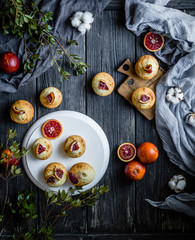 Muffins with blood oranges on a white wooden stand with green leaves, cloth and wooden desk on a black background