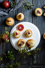 Muffins with blood oranges on a white wooden stand with green leaves, cloth and wooden desk on a black background