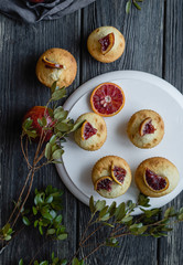Muffins with blood oranges on a white wooden stand with green leaves, cloth and wooden desk on a black background