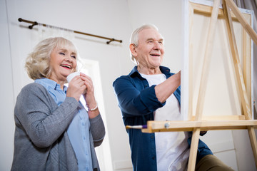 Happy senior couple painting picture on easel at home