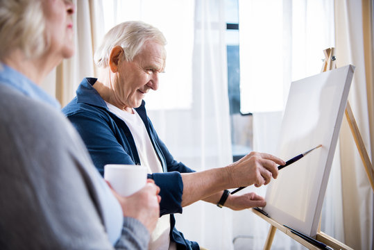 Senior woman holding cup near smiling man painting picture on easel