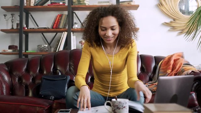 young latin curly woman speak with friend by telephone mobile using handsfree earpieces and look through magazine simultaneously while sitting and resting at home with cup of tea during sunny day