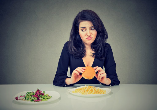 Junk food vs healthy food. Hesitant woman with cheeseburger looking at vegetables salad
