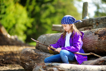 Cute little girl sitting on tree logs using a pocket knife to whittle a hiking stick © MNStudio