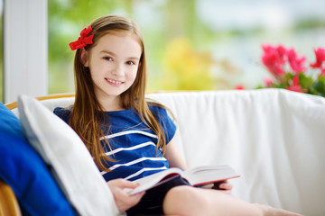 Adorable little girl reading a book in white living room on beautiful summer day