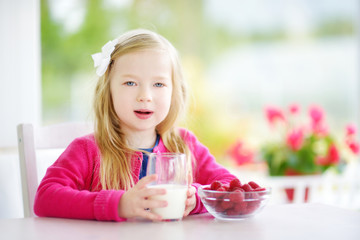 Pretty little girl eating raspberries and drinking milk at home. Cute child enjoying her healthy fresh fruits and berries.