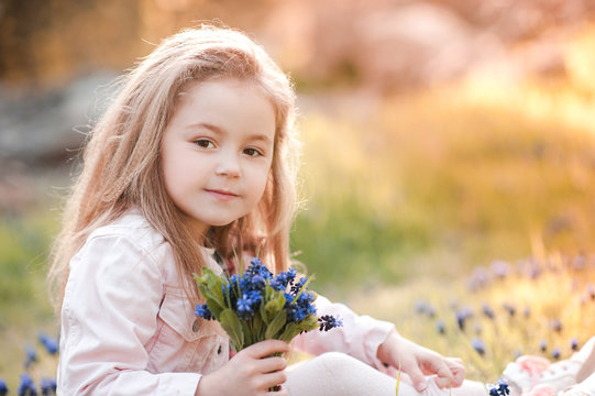 Smiling Baby Girl 4-5 Year Old Holding Blue Flowers Outdoors. Looking At Camera. Posing Outdoors. Summer Time.