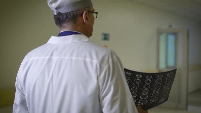 A neurosurgeon physician who is dressed in a white robe walks along the corridor of the clinic and holds the results of brain tamography