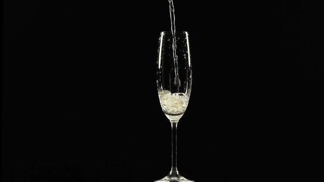 Sparkling yellow water is pouring into wineglass standing on black background in slowmotion