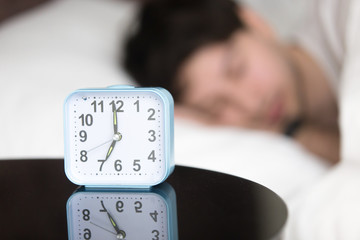 Young guy peacefully sleeping in white cozy bed in the morning in the background. Focus on the alarm clock standing on the table near the bed, telling early time. Before waking up