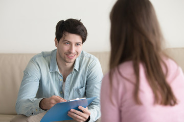 Young smiling man sitting on couch opposite woman, holding clipboard, talking with patient or asking personal information. Visiting psychologist, filling up form or questionnaire, informal interview