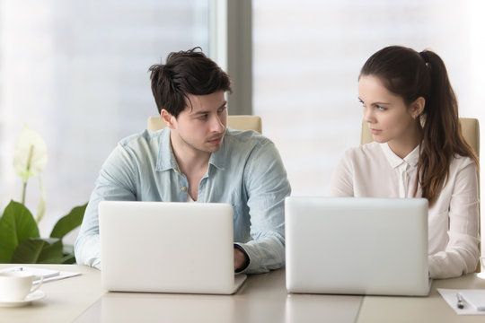 Competition and rivalry between office workers. Male and female colleagues sitting together at conference table using laptops looking in the eye, dislike each other, feeling jealous, being envious