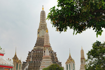 Fototapeta premium Stupa encrusted with coloured faience in Temple of Dawn, Wat Arun, Bangkok, Thailand. Ancient Thai temple surrounded by tropical trees against the cloudy sky
