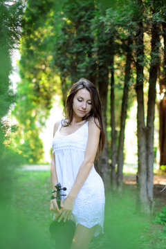 Красивая девушка в коротком белом платье стоит со скрипкой в лесу и смотрит вниз 
