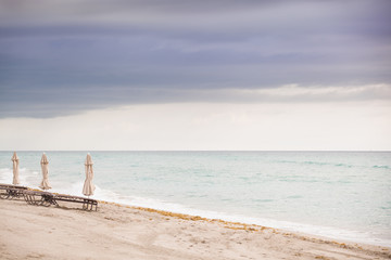 Beach in Miami, FL. Folded umbrellas on the sand beach at the ocean. Empty beach. Sea shore. Early morning. Clouds background.