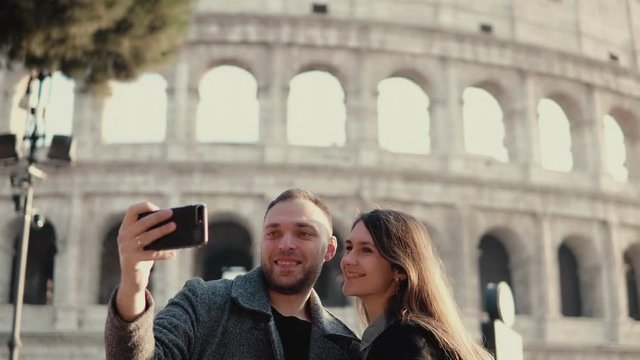 Young attractive woman and man standing near the Colosseum in Rome, Italy. Couple takes the selfie photo on smartphone.
