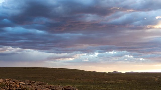A slow pan and tilt timelapse of a vast open landscape with rocky foreground while following stormy magenta clouds and rain falling in the distance against a dark, dramatic sky