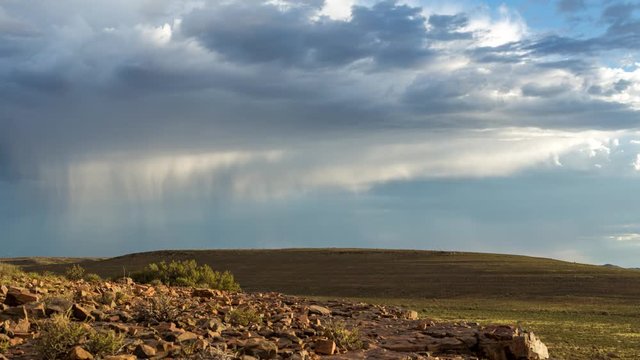 A slow panning timelapse of a vast open landscape with rocky foreground while following stormy clouds and rain falling against a dark, dramatic sky