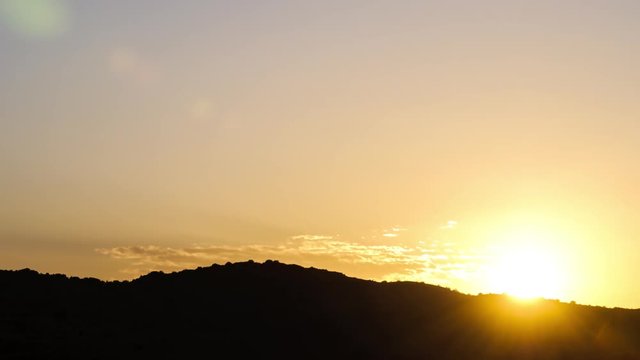 Static sunrise timelapse with orange and yellow glow, scattered clouds against a silhouetted mountain on the horizon in the foreground with sun flare as the sun rises above the mountain available on request.
