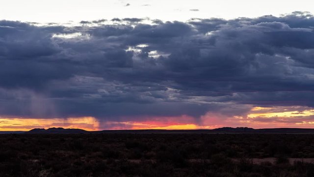 A static timelapse with dark and dramatic deep blue stormy clouds at sunset with golden and magenta  glow and patches of rain drizzling over the landscape, anywhere
