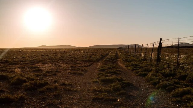 A linear timelaspe at sunset with sun flare of a typical Karoo farm landscape framed by a fence with a dirt road leading you towards the distant mountains and hills