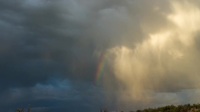 A static timelapse slowly tilting up from a rocky landscape foreground to a dramatic stormy sky with a colourful rainbow against dark blue and bright clouds while rain is pouring down