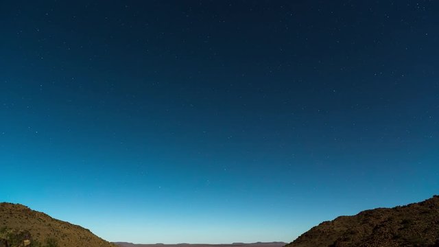 A static timelapse tilting down at night over vast mountain landscape with wide open moonlit plains below and a sky filled with stars against a blue sky