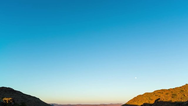 A static timelapse tilting up towards a bright blue sky at sunrise over vast mountain landscape with wide open plains below as the moon sets