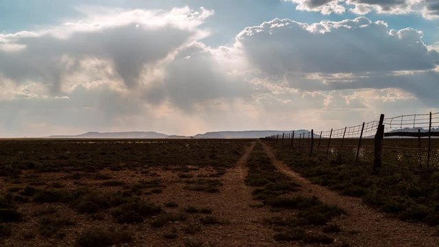 A linear timelaspe at sunset with sun flare of a typical Karoo farm landscape with scattered cumulous clouds looking like rain, framed by a fence with a dirt road leading you towards the distant mountains and hills