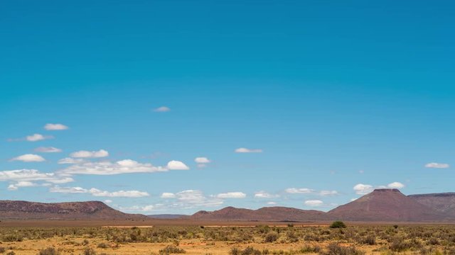 A static daytime timelapse of an arid (semi-desert) landscape in the Karoo, South Africa with shrubs and hills (mountains) against a blue sky with scattered cumulous clouds