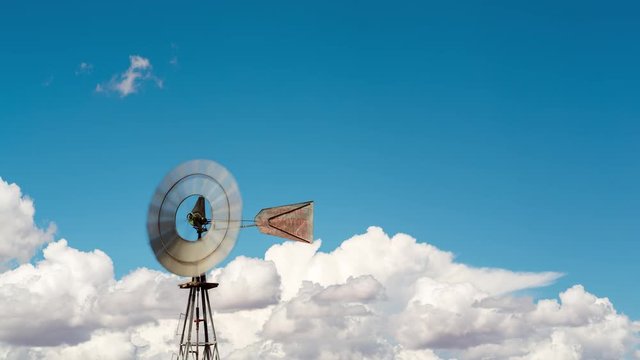 A static medium daytime timelaspe shot of a windmill frantically blowing in the wind against a bright blue sky with cumulous clouds, front view