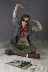 Attractive young female artist with dreadlocks sitting on a floor, holds brushes, there are tubes with paints nearby