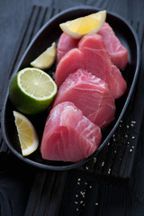 Frying pan with raw sliced tuna fillet with lemon and lime, vertical shot on a black wooden background, close-up