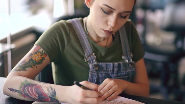 Closeup of young attractive red haired woman tattoo artist sitting at table and creating sketch for tattooing in studio indoors