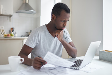 Young serious african american man sitting at kitchen table in front of open laptop, concentrated...