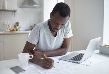 Portrait of dark-skinned man, sitting at kitchen table, filling application form, writting...
