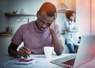 Sad african american man sitting with laptop at kitchen table, looking at bills, counting expenses,...