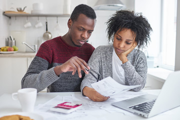 Young African American wife and husband sitting at home with laptop, calculator and papers doing...