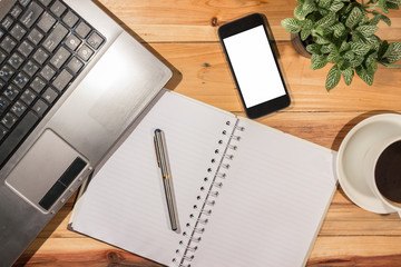 Top view of wooden desktop with laptop, blank mobile phone, spiral notepad, decorative plant and coffee.