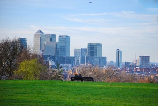 Couple sitting down on a bench on hill looking at the London Canary Wharf Skyline in Greenwich