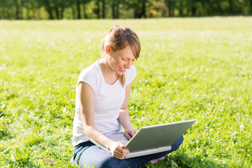 Beautiful smiling young woman sitting on a meadow and using her laptop