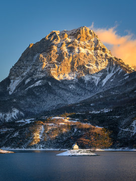 Saint Michel Chapel across Serre Poncon Lake in the Southern French Alps. The sunlit peak of Grand Morgon rises above Saint Michel Bay at sunset after a snow storm.