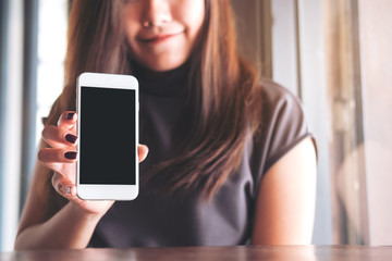 Mockup image of a beautiful woman with smiley face holding and showing white mobile phone with blank black screen  in vintage wooden cafe