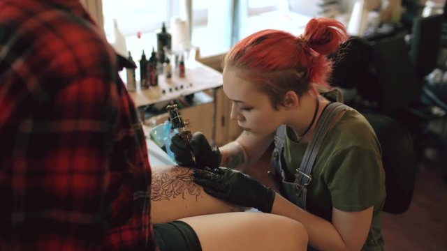 Red haired girl tattoo artist tattooing picture on lef of young girl client in studio