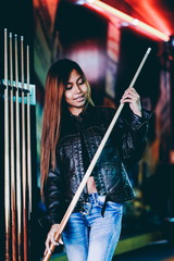 Young beautiful girl wearing leather jacket in a billiard club, with cue stick preparing for the game