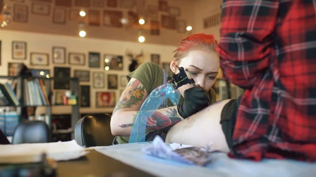 Tilt up shoot of young red haired woman tattoo artist tattooing picture on leg of young girl client in studio