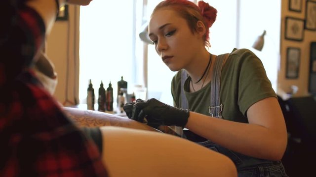 Young red haired woman tattoo artist tattooing picture on leg of young girl client in studio