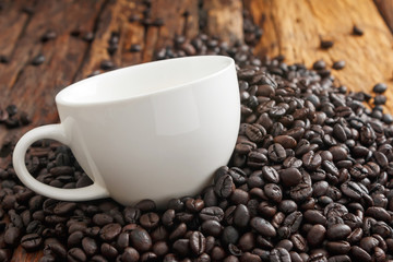 Close up shot of white ceramic cup on pile of dark roasted coffee bean and wooden floor with copy space