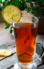 High glass cup of black tea with a slice of dried orange on a bright wooden rustic background  and foliage.