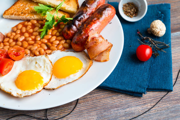 Closeup of full english breakfast in whte plate on wooden table with blue linen napkin.